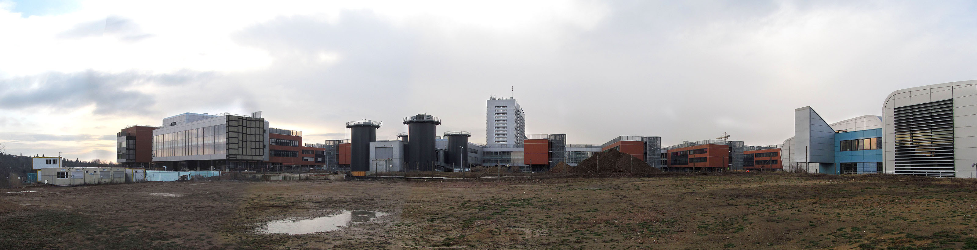 A tohle je lednové panorama kampusu od severu. Na tomto místě, v nedávné minulosti skládce, budou zanedlouho stát další oranžové pavilony a třetí dvouposchoďový koridor. Vlevo je dobře vidět jeho první část, zaslepená sádrokartonem pospojovaným černou hmotou, vpravo obdobná, uží záslepka vycházející z modré budovy FSpS. Třetím, prozatím slepě ukončeným tubusem, který spojí tuto část kampusu přes most s jeho hlavním prostorem, je garážovitě vyhlížející budova mezi černými věžemi (v současnosti je uvnitř kavárna).