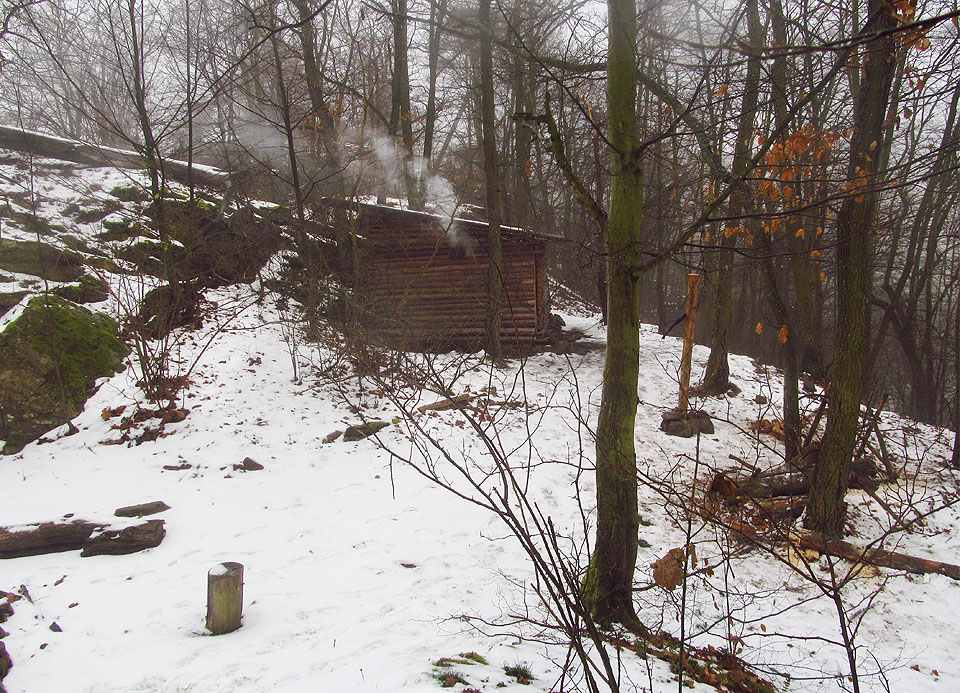 Tábořiště na Prasklém džbánu v zimě. Osiřelé kmeny okolo ohniště, němý kůl na hody mačetou či sekerou a pouze tlumeně znějící hlasy ze srubu, kde u kamen přežívají jediné živé duše.