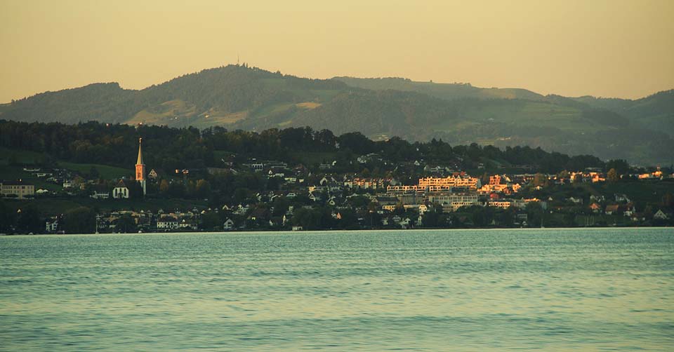 Poslední paprsky osvětlují pobřeží Curyšského jezera. Voda je průzračná, na břehu spousty plavců, restaurace, pláže kultivované i přírodní a železnice prakticky skoro na břehu.