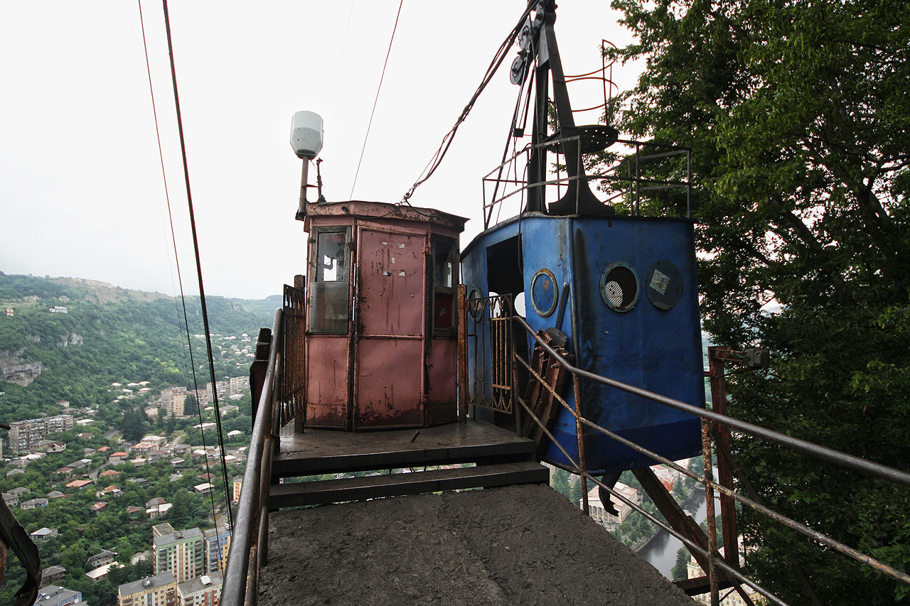 Modré kabinky nahoře „přistávají“ u betonového mola vytrčeného do prostoru nad skálou obzvláště akrofobickým způsobem. V růžové boudě sedí paní lanovkářka, která po druhém zazvonění zvonku ze spodní stanice spouští motor.