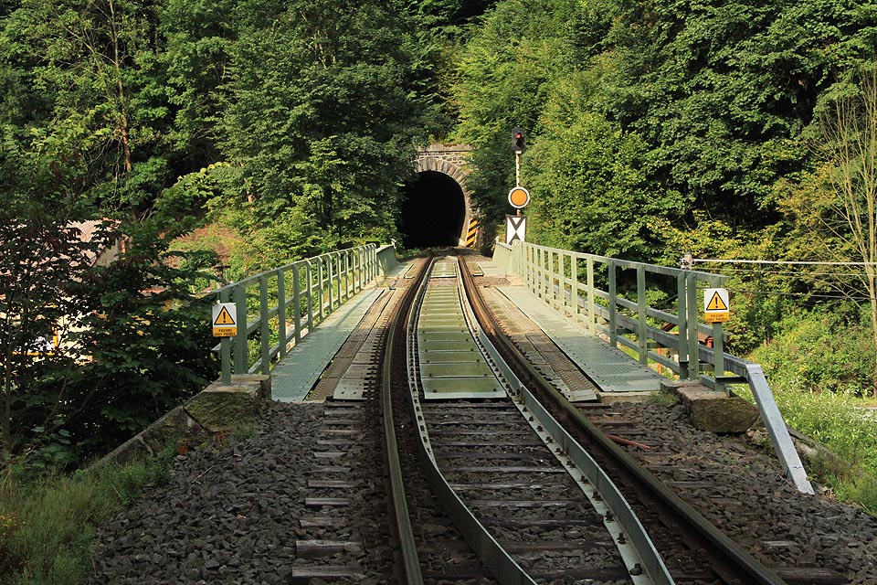 Příjezd k Bečovskému tunelu od severu, přes mosty nad řekou Teplou a silnicí č. 20 (E49). V srpnu 2015 probíhala výměna původních mechanických návěstidel ve stanici <a href=/Tema/Be%C4%8Dov%20nad%20Teplou%20%28%C5%BEst%29>Bečov nad Teplou</a> a na předvěsti byla instalována nepřenosná pevná návěst výstraha.