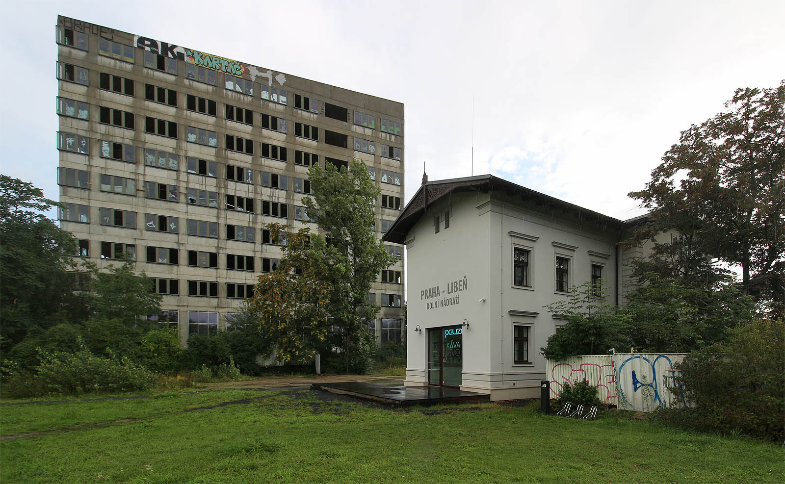 Kolejiště stanice <a href="/Tema/Praha-Liběň dolní nádraží (žst)">Praha-Liběň dolní nádraží</a> se nacházelo severně od staniční budovy (na snímku blíže k fotografovi), mezi histroickou budovou a současným betonovým monstrem se nacházelo několik kolejí vleček a rozsáhlý areál skladů, ze kterých nezůstalo nic. Koleje odsud ale tak úplně nezmizely: přímo pod cípem centra Nová Palmovka prochází od roku 1990 tunely metra B.