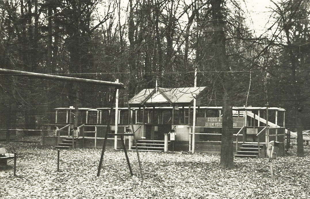 Hlavní stanice přerovské pionýrské železnice se soupravou vozů bez lokomotivy (v pozadí skluzavka na dětském hřišti). Štěstí a mír (a vlaky) dětem všech národů! <br>Datum snímku je pouze přibližné, autorem je Vladimír Mokošín.