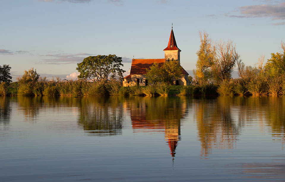 Neogotický kostelík sálá harmonií vesnického podvečera. Na ostrov doprostřed vodní nádrže nepatří - jako by sem byl přenesen omylem nějakou obří silou úplně odjinud.
