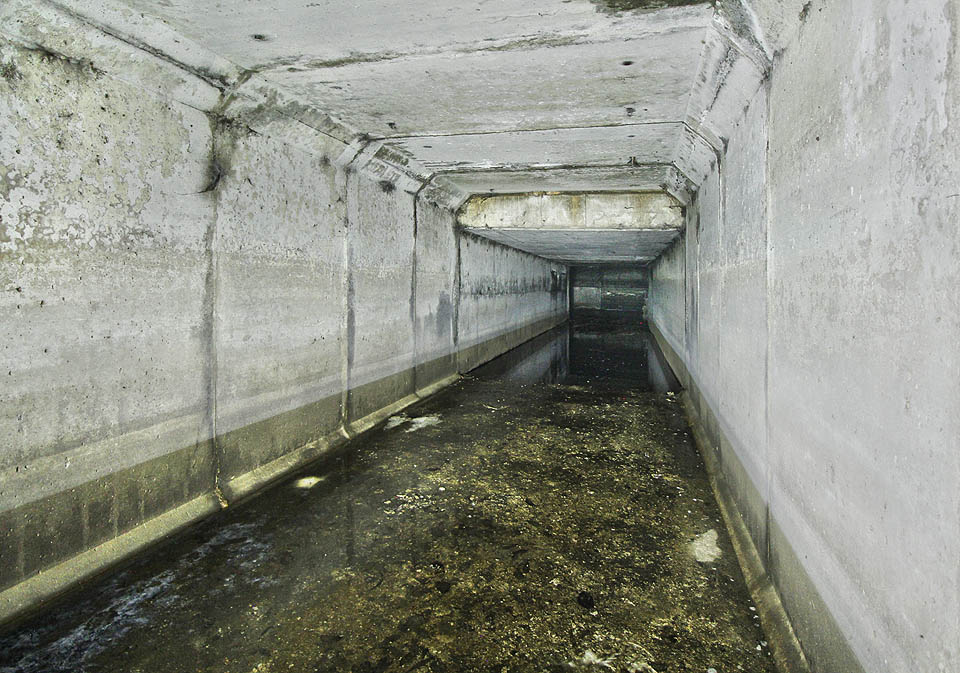 Pokračování spojovací chodby směrem k odlehčovací komoře OKB04. Dobře patrné je snížení stropu pod železniční vlečkou a v dálce zahrazená stěna koryta kmenové stoky B.