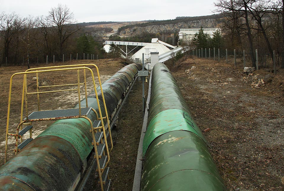 Začátek dvojice dopravníkových pásů je u krytého (dříve však volného) kuželového zásobníku suroviny na okraji lomu v Mokré. To, co vypadá jako pomačkané roury, jsou plechové kryty horního pásu.