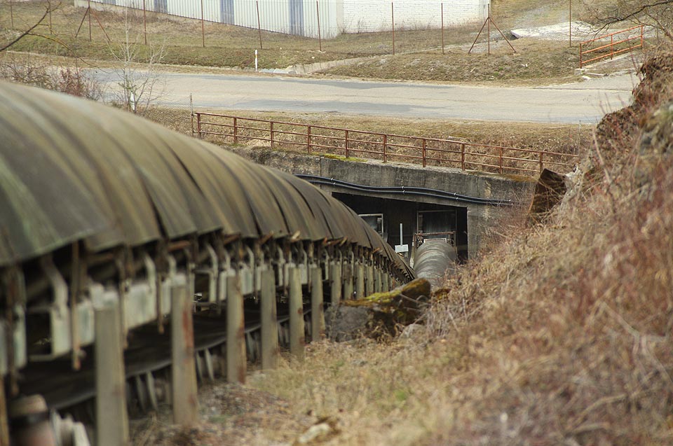 Ze zásobníku kameniva pod silnicí vedou oba transportní pásy pod zemí. Vypadají jako obrovská lesklá stonožka, která vylézá ze své škvíry pod kamenem.