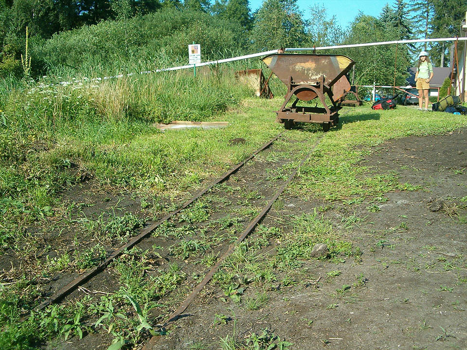 Odstavná kolej s typickým vozíkem pro manuální přepravu rašeliny. V pozadí další vozík ve vyklopené poloze a odvážná školačka, toužící po svezení.