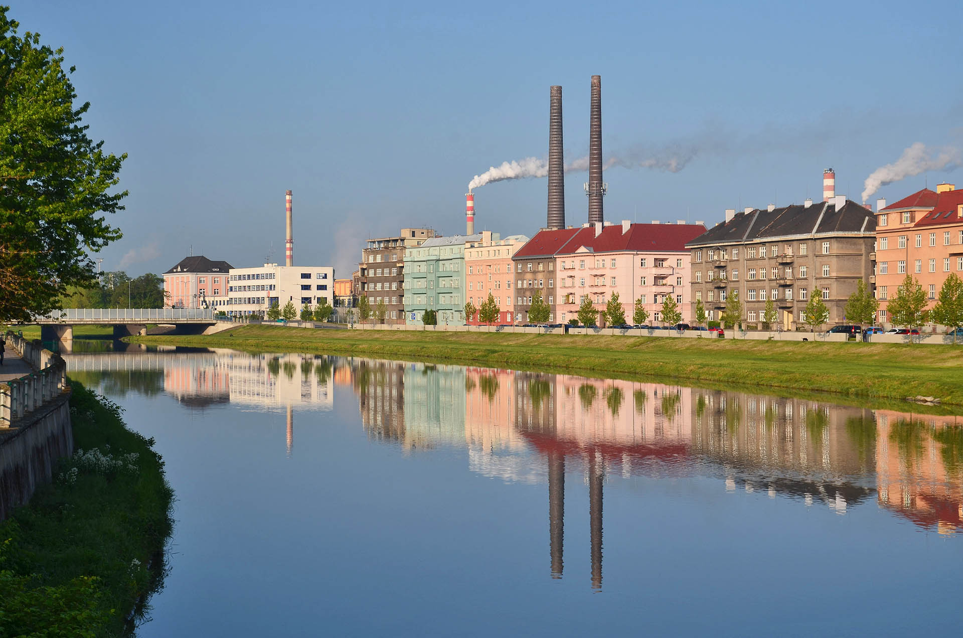 Pohled na komíny elektrárny z nábřeží Protifašistických bojovníků přes protější nábřeží Dr. Edvarda Beneše a mírně zčeřenou hladinu Bečvy.