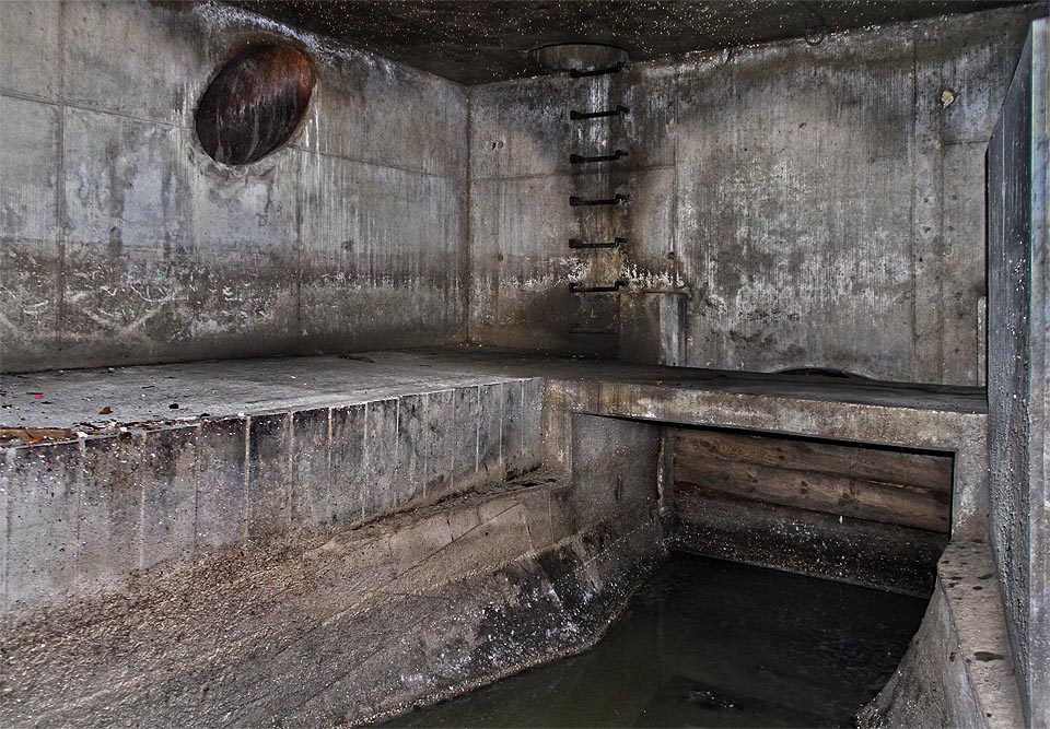 Pohled z pravé části komory k jihovýchodu. Dřevěným pažením je omezen vtok do užšího profilu stoky C01 vedoucí podle náhonu. Pochopit účel kovové roury ostře spadající od stropu po zvláštním úhlem kamsi do temné hlubiny, se nám ovšem nepodařilo.