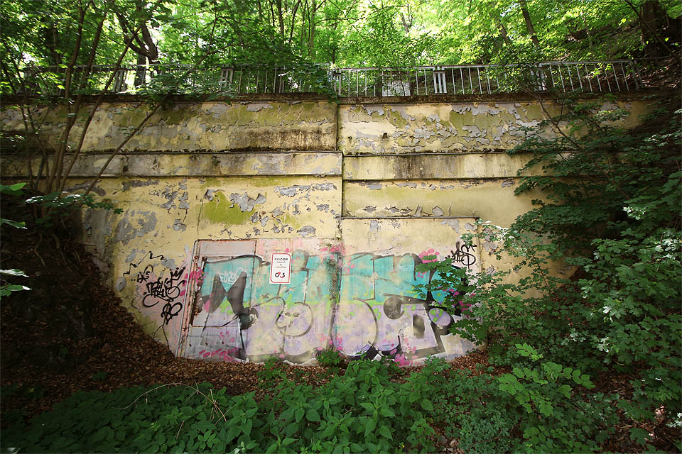 Hlavní vstup do nejdůležitějšího z adamovských krytů zrovna honosně nevypadá. Obklopen zelení a potřísněn hloupými sprejery mlčenlivě kryje jícen podzemní hlubiny.